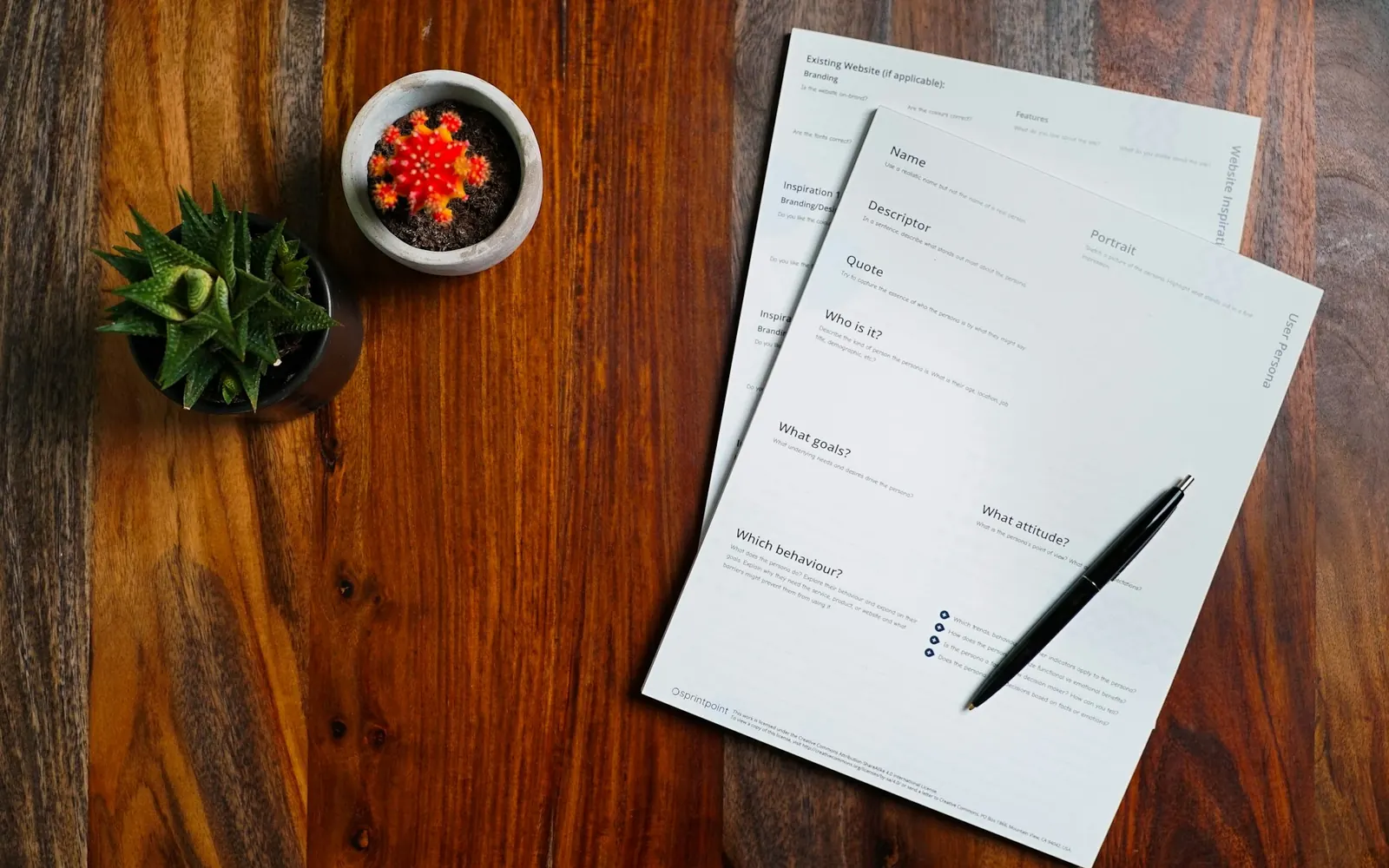 Illustrative photo: printed role specification pages and a pen on a wooden desk. Not a sample of the Job Description & Hiring Pack deliverable.